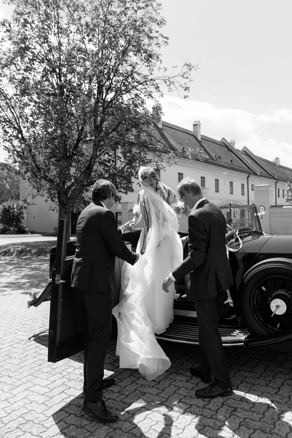 A vintage black and white wedding scene showing a bride and groom near a classic car in front of residential buildings.