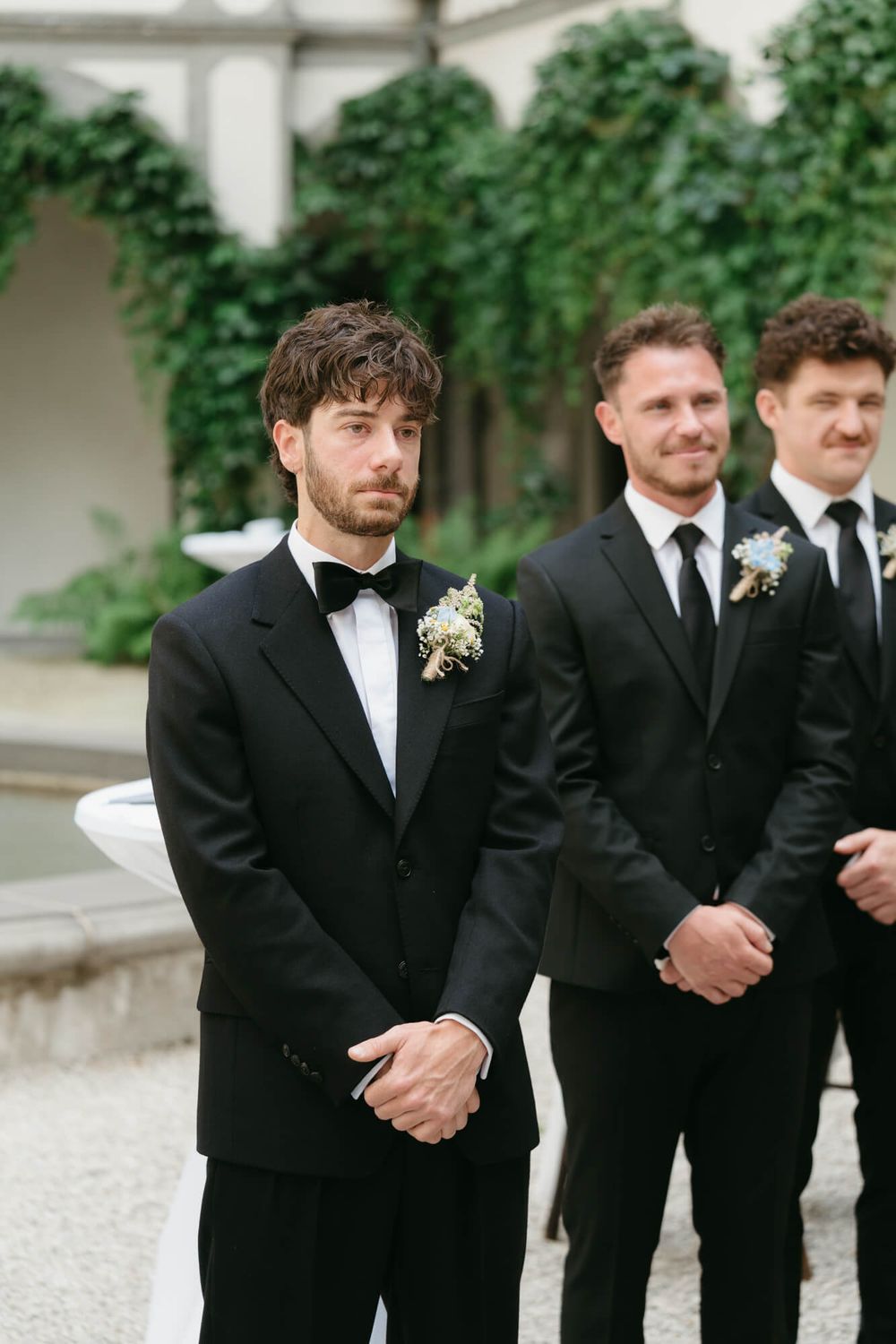 A line of groomsmen in black tuxedos and ties stand together at an outdoor wedding ceremony.