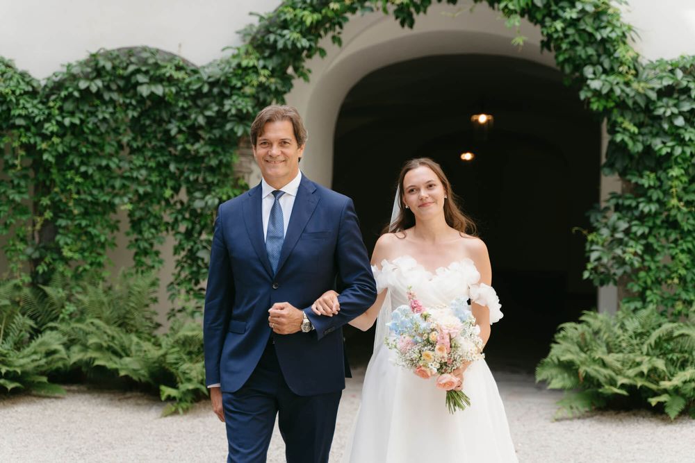 A bride and groom walk arm in arm through a garden archway during their wedding ceremony.