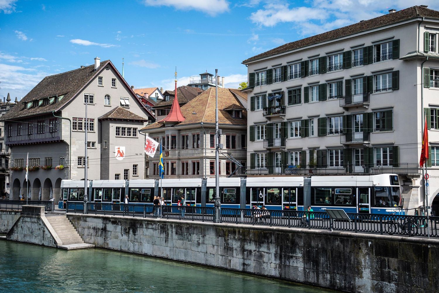 Historic buildings line a scenic canal waterfront in a quaint European town under blue skies.
