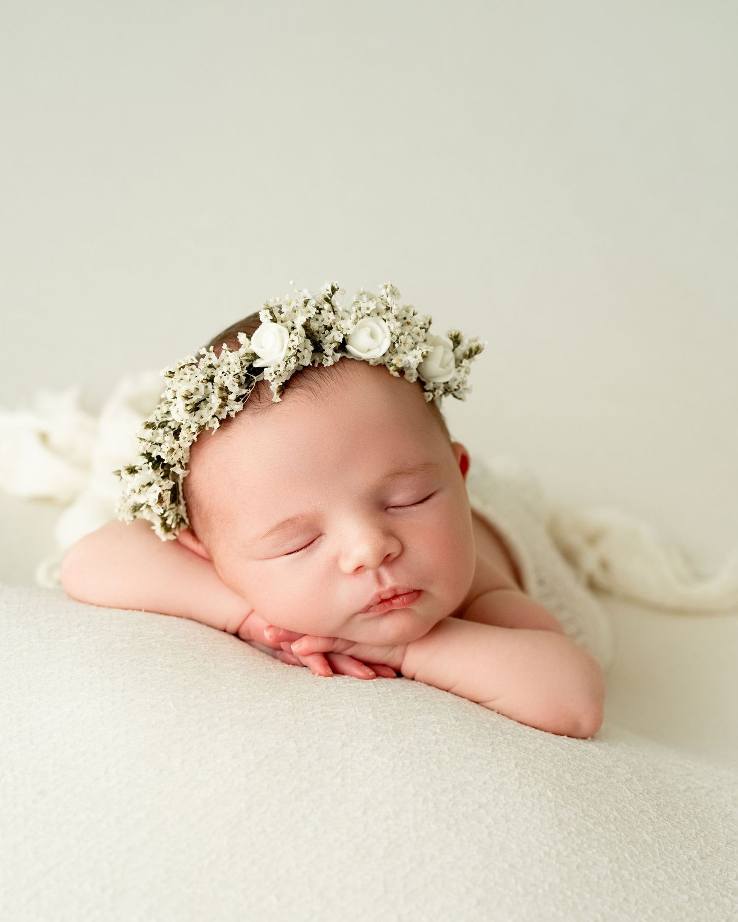 Sleeping newborn baby wearing delicate floral crown rests peacefully on white blanket in professional photography setup.