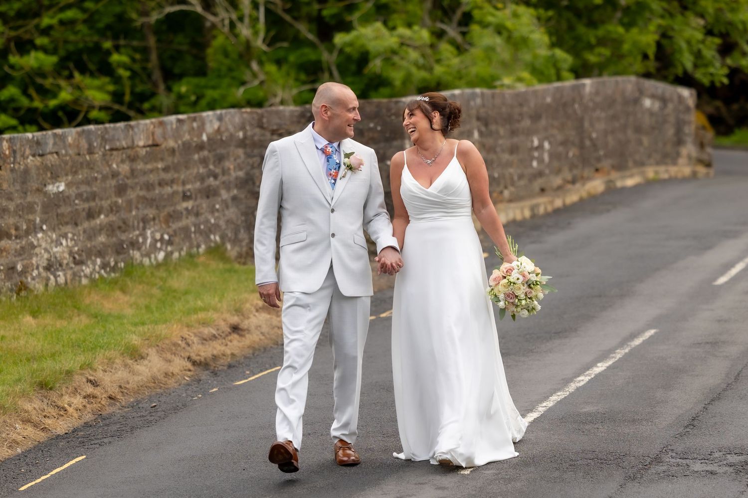 A couple in white wedding attire walks together on a paved road surrounded by stone walls and greenery.