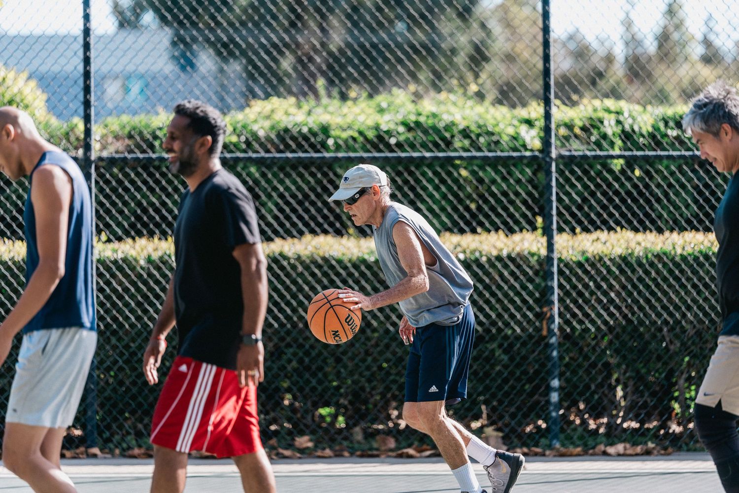 Basketball players compete on an outdoor court during a pickup game with chain link fence in background.