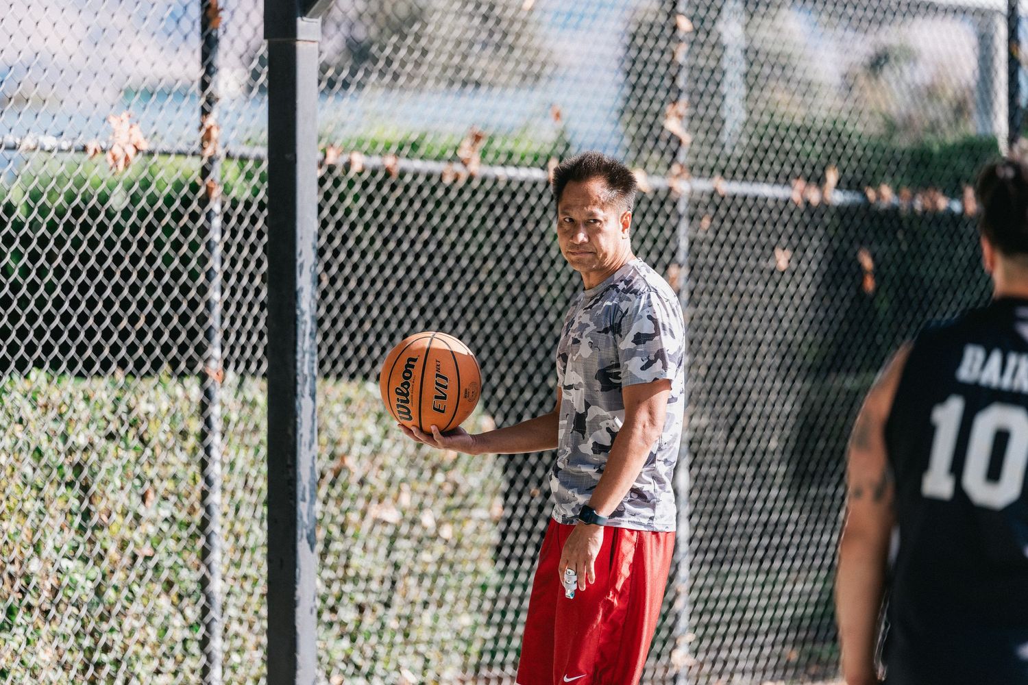 Basketball player in camouflage shirt and red shorts holding ball on outdoor court.
