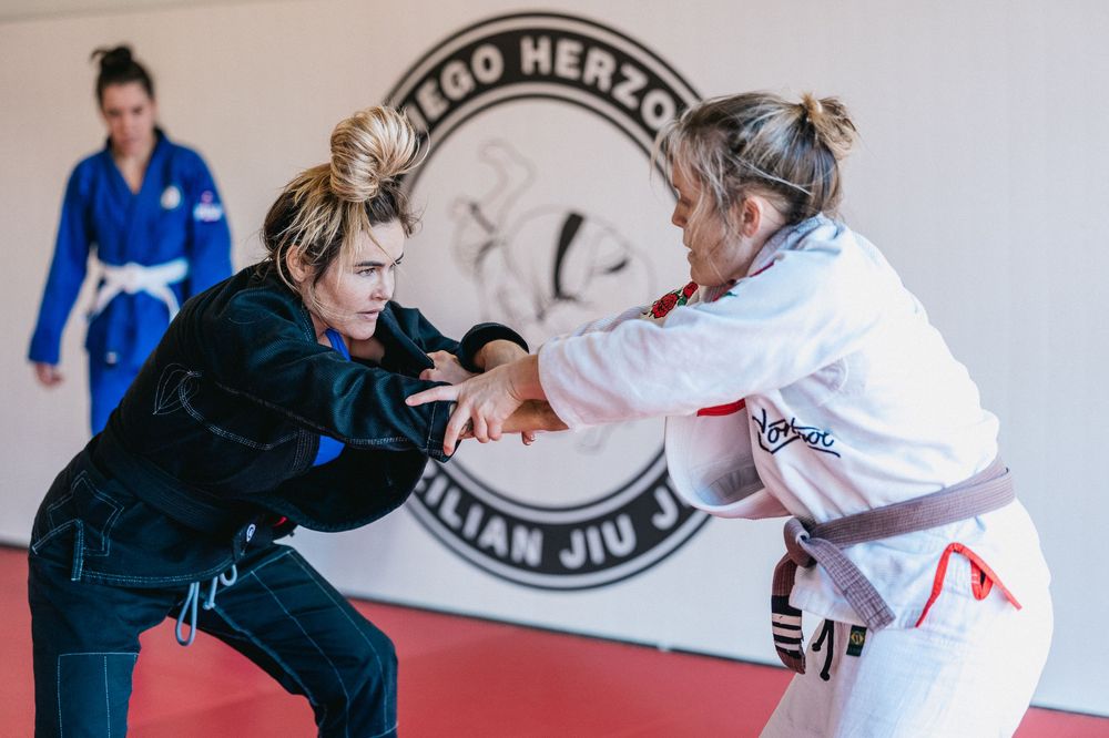 Two martial artists practicing jiu-jitsu techniques on a training mat with a gym logo visible in the background.