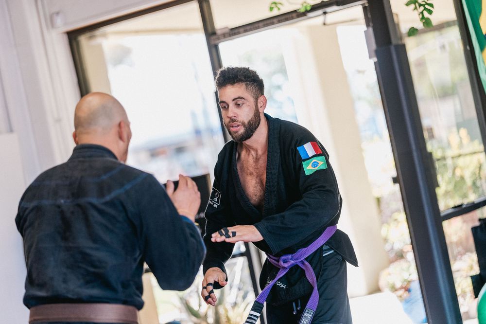 Two martial artists practicing Brazilian Jiu-Jitsu techniques in a training session, one wearing a black gi with purple belt.