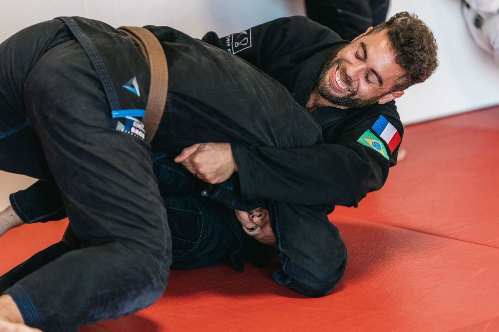 Two martial artists demonstrate a ground fighting technique on red mats during Brazilian Jiu-Jitsu training.