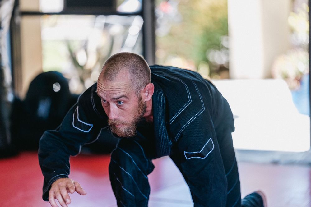 A martial artist in a black gi crouches in a ready position on a training mat.