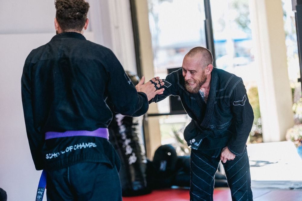 Two people practicing martial arts techniques in a training studio with bright windows in the background.