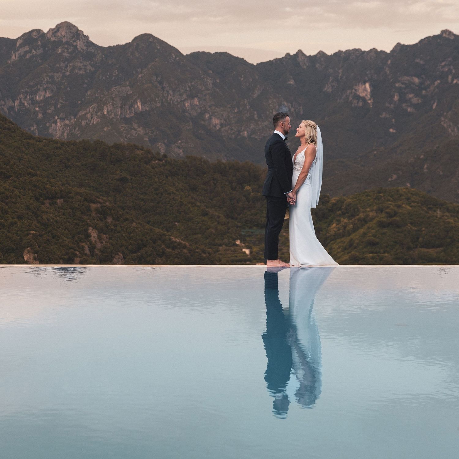 A couple shares an intimate moment at sunset against a backdrop of rugged mountains and a reflective lake.