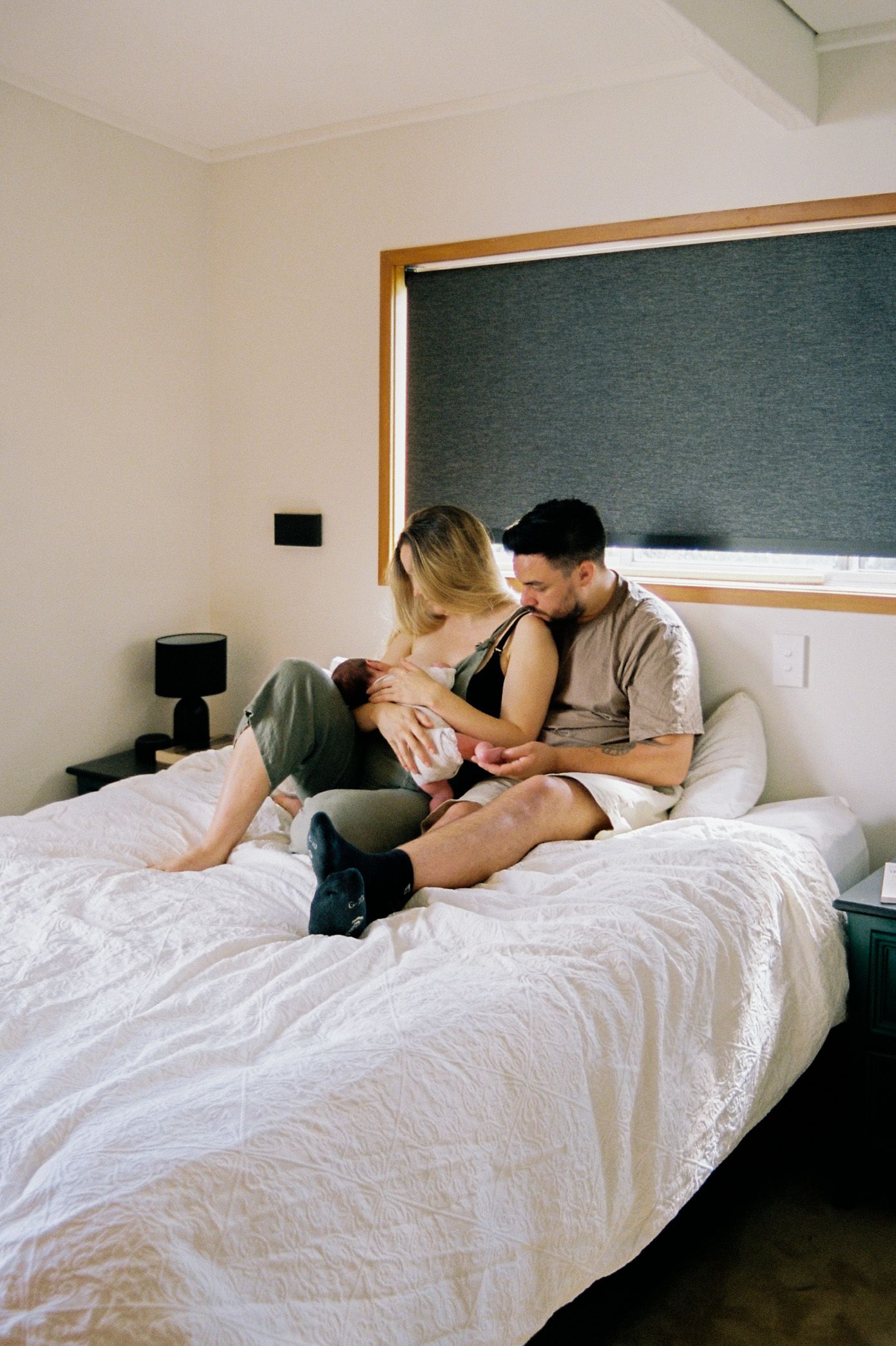 A couple sits together on a white bed in a cozy bedroom with a dark green accent wall.