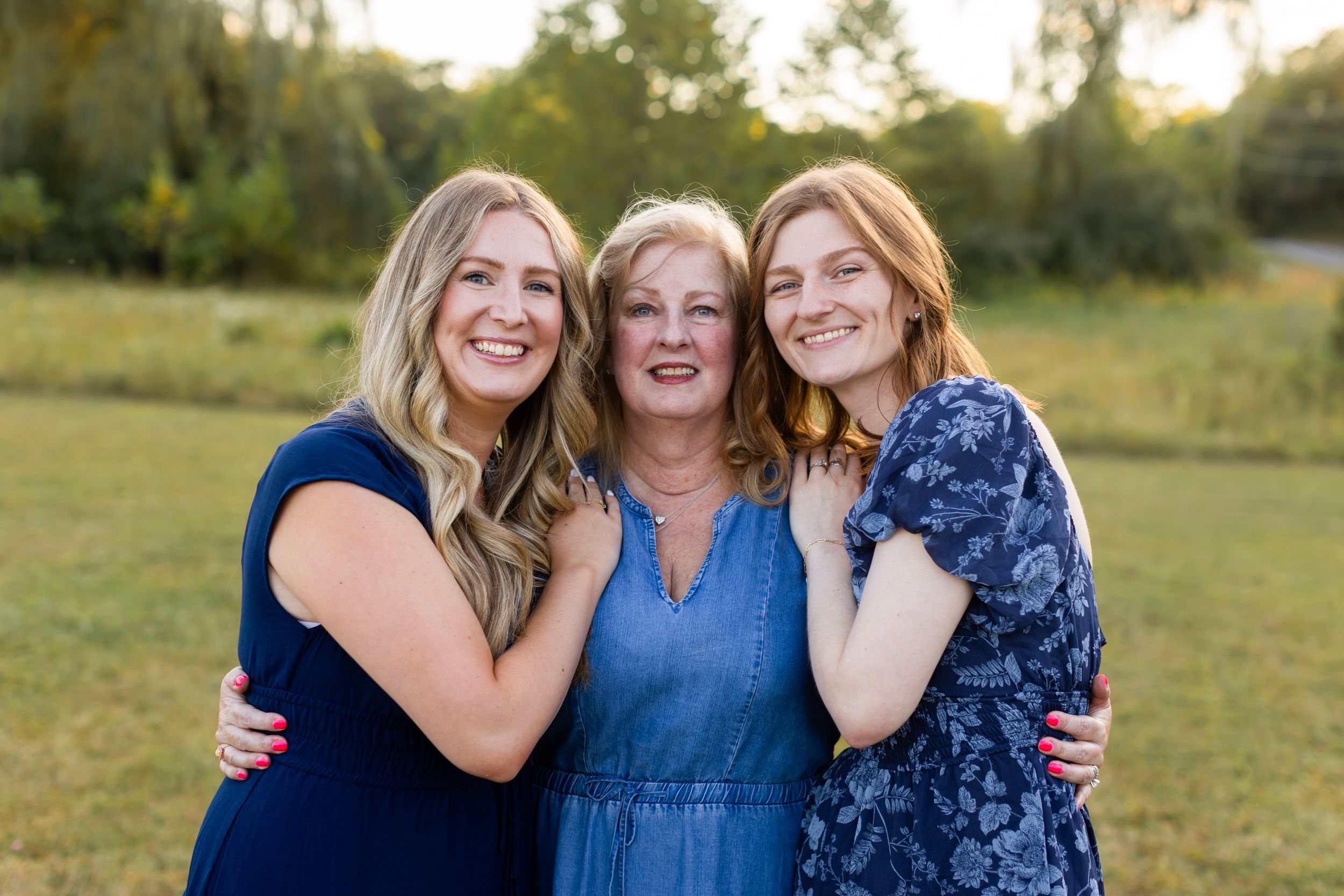 Three women in coordinating blue dresses embrace and smile together in a sunny outdoor setting. NJ extended family photographer