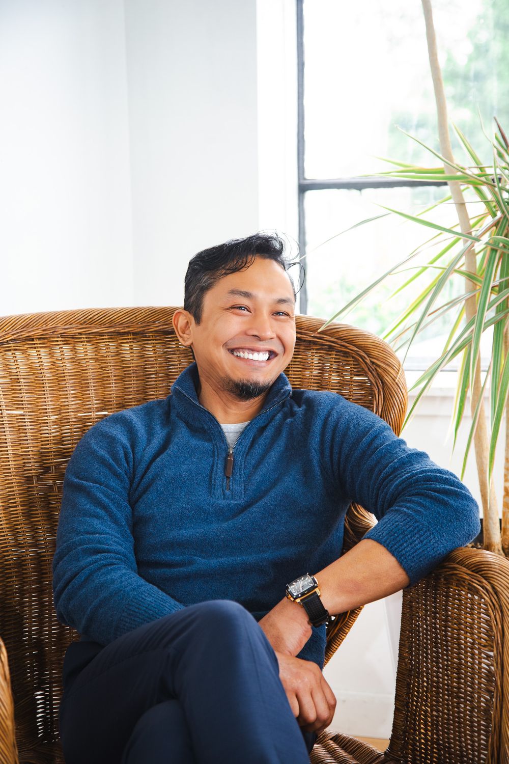 Smiling man in blue sweater sitting relaxed in wicker chair near window with tropical plant.