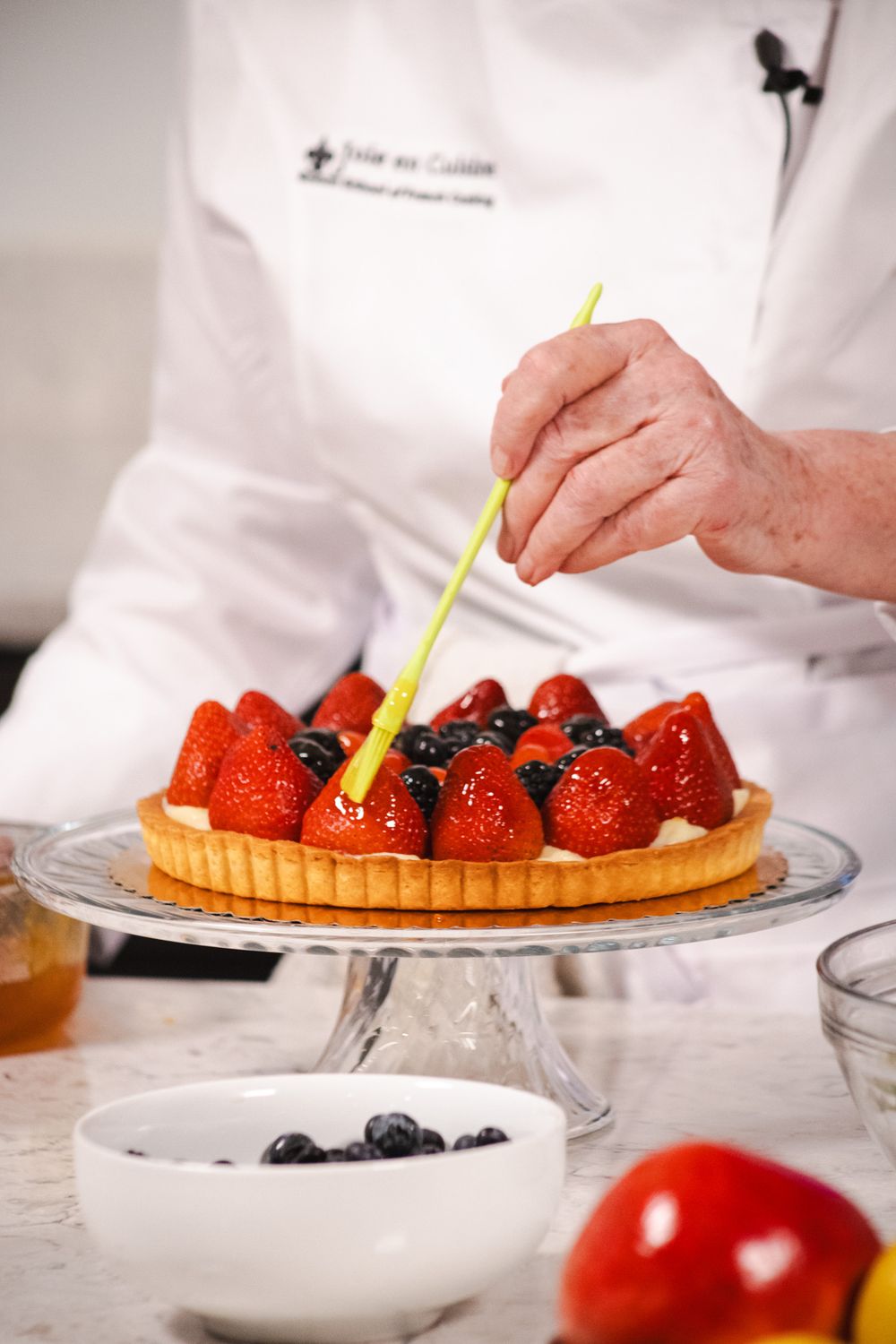 Chef arranges fresh berries on a tart with tweezers for precise presentation on a glass cake stand.