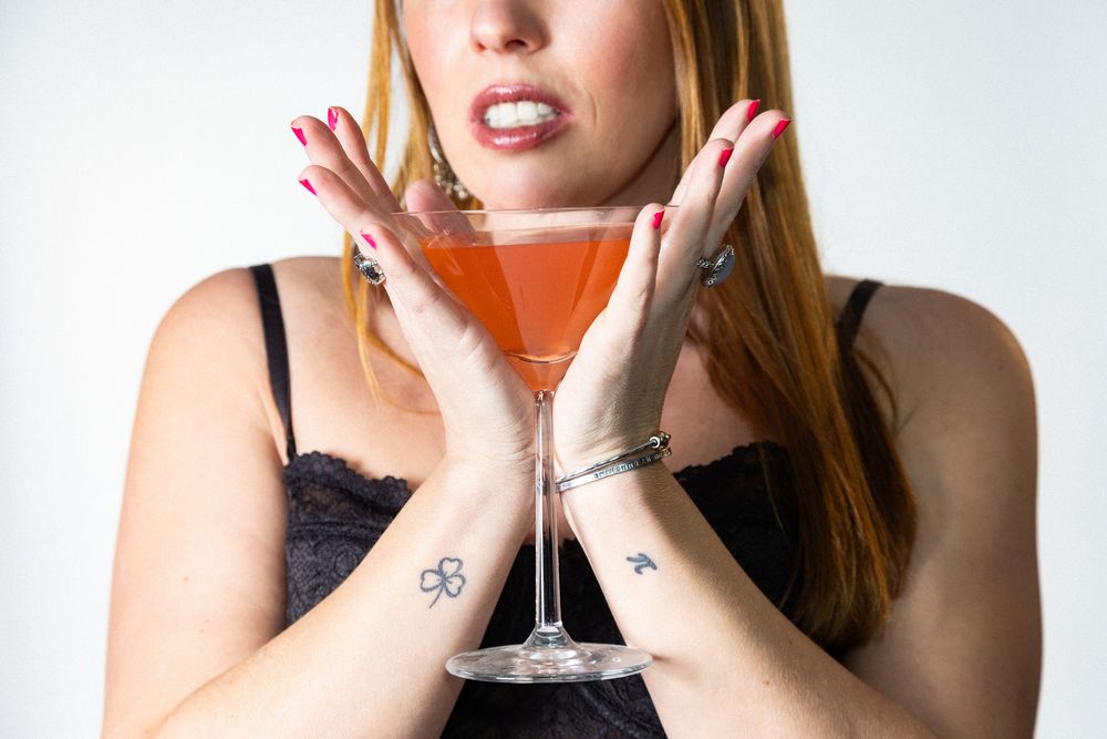 A person holds up a martini glass with a pink cocktail while smiling against a white background.