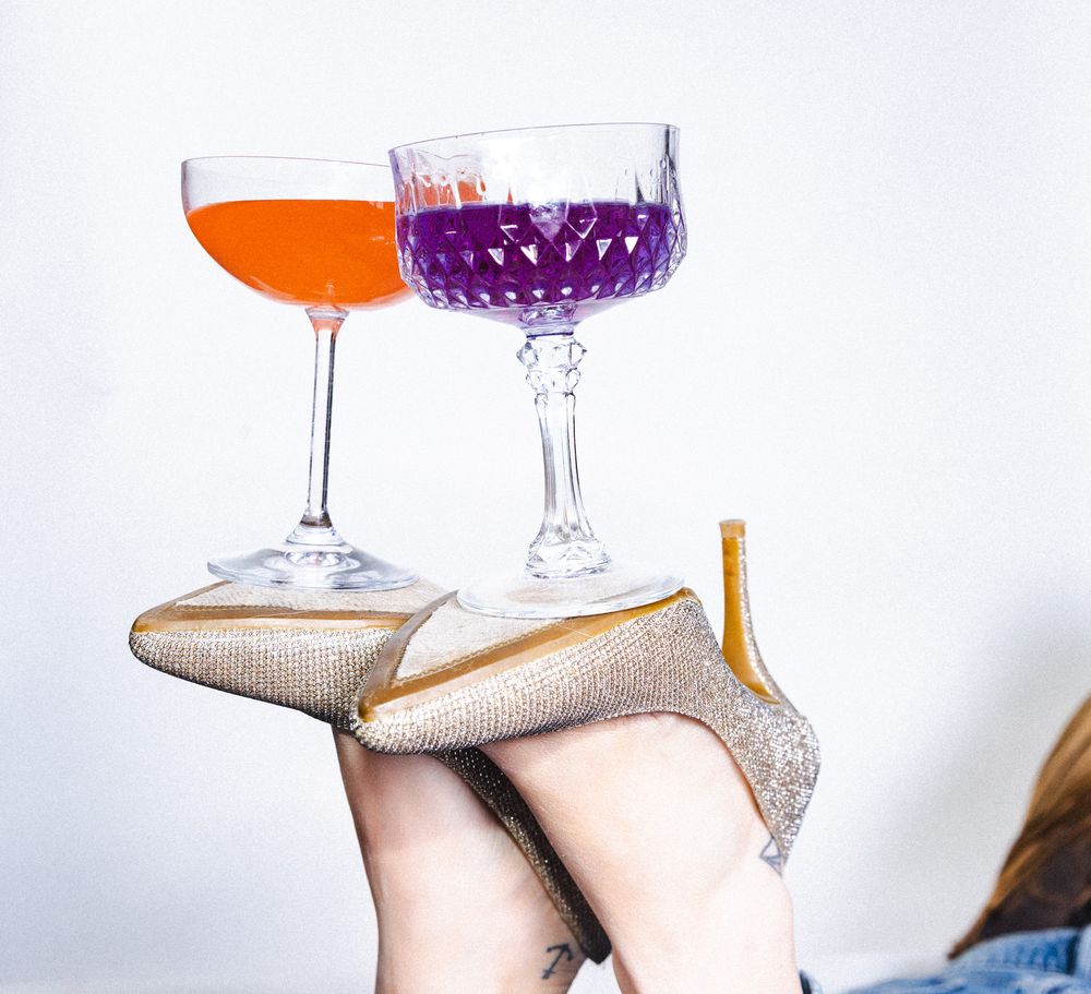 Two cocktail glasses balanced on silver high heel shoes against a white background.
