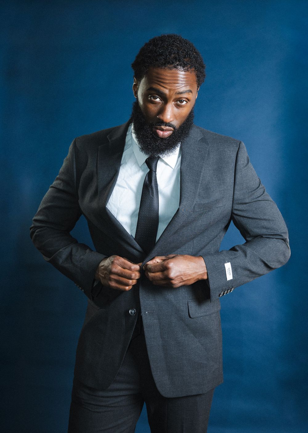 Professional portrait series showing a person in a grey suit adjusting their jacket against a deep blue studio backdrop.