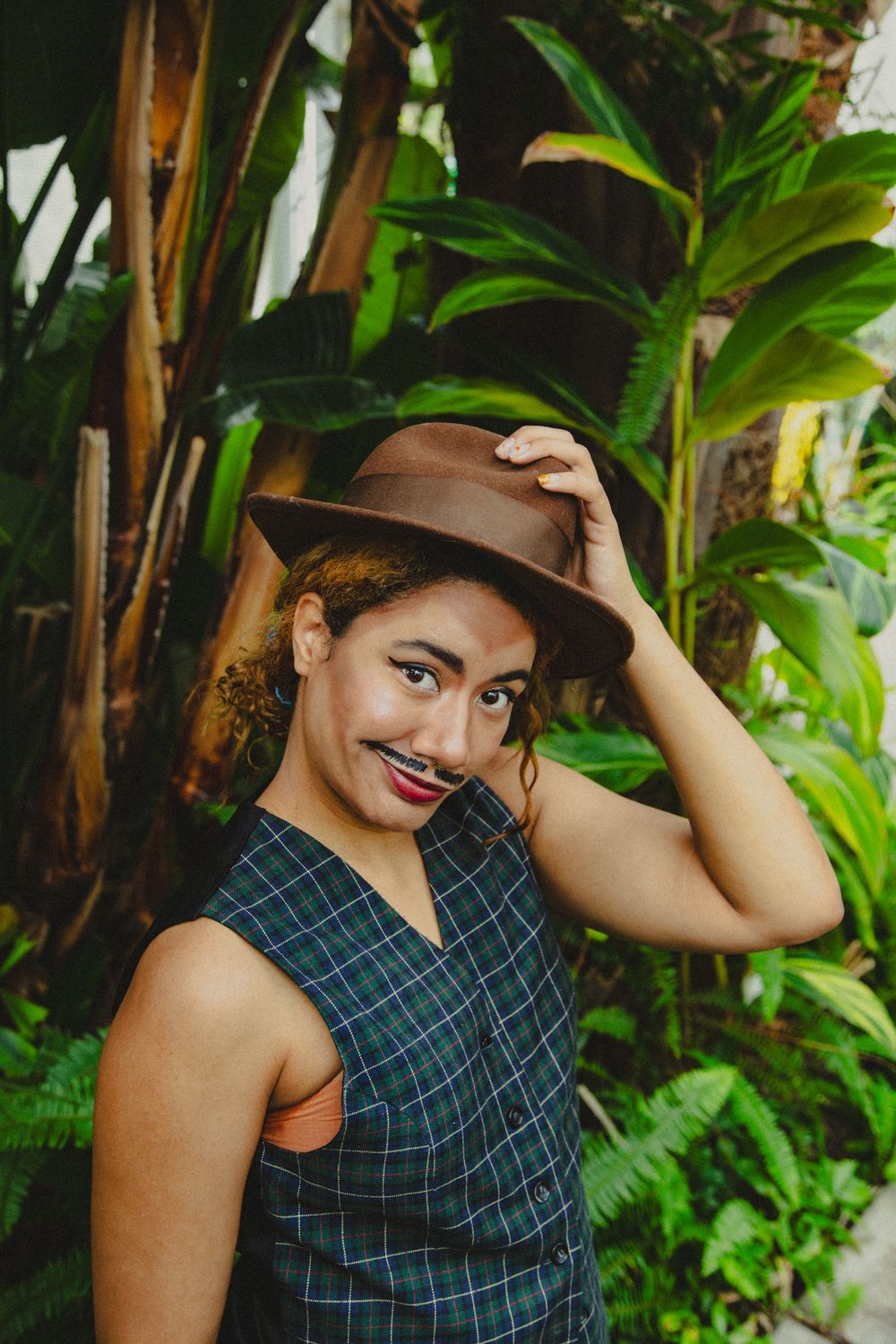 Woman in plaid vest and brown fedora hat poses playfully with fake mustache among tropical green plants.