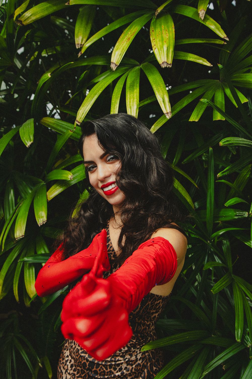 Smiling woman in leopard print dress and red gloves posing in front of lush tropical green foliage.