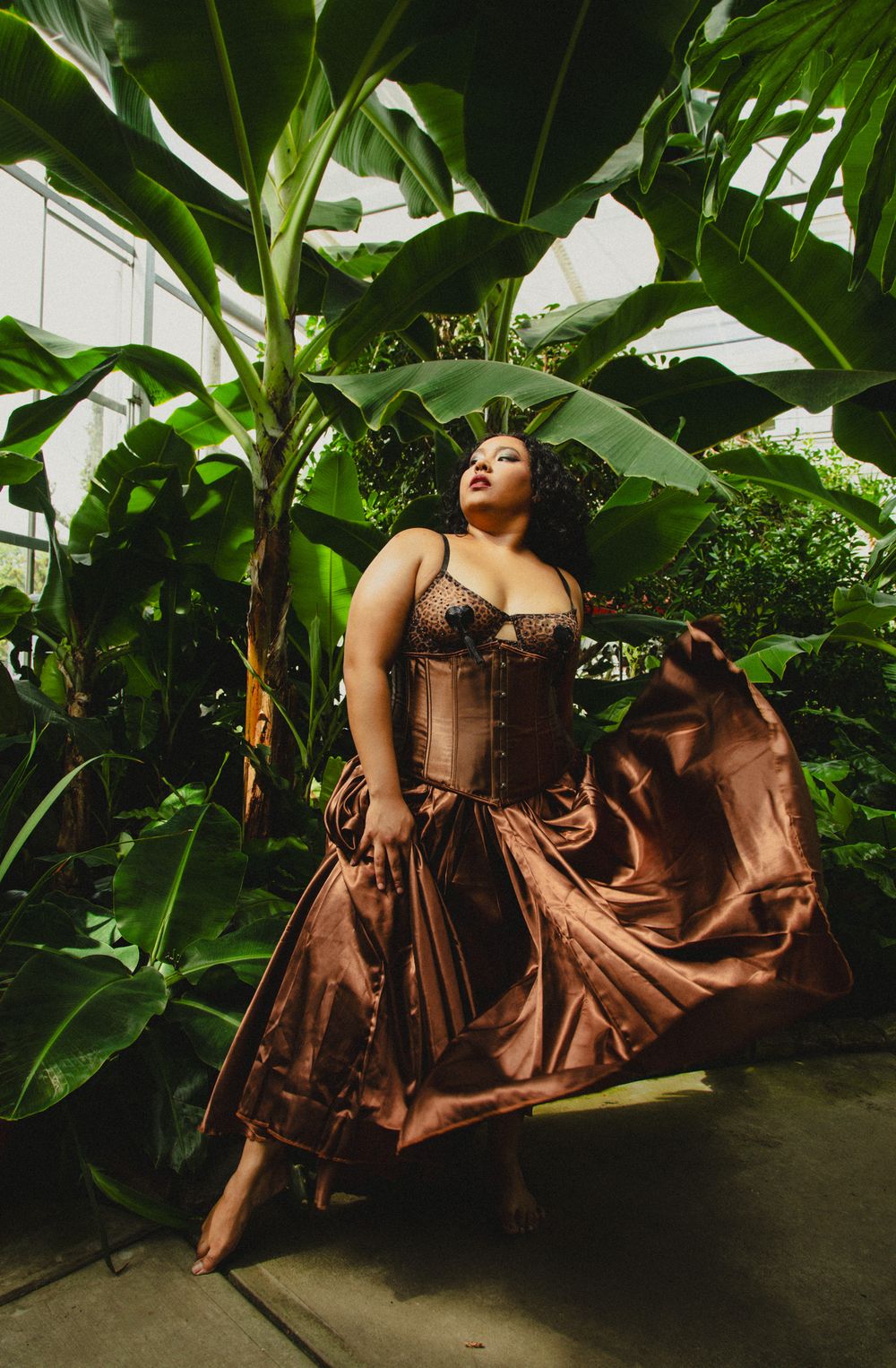 Woman in bronze corset gown poses gracefully among lush tropical plants in a greenhouse setting.
