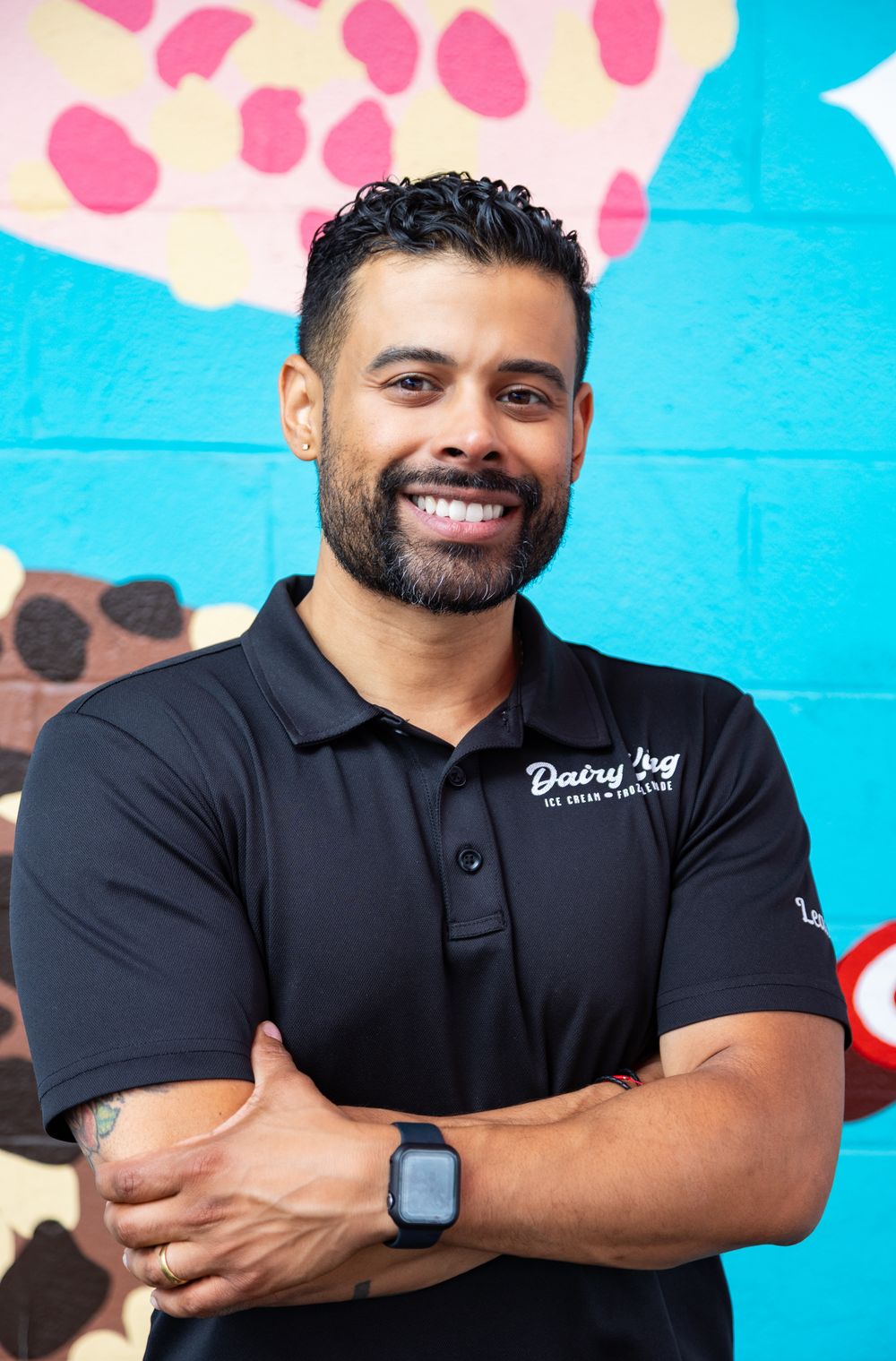 Smiling man in a black Dairy Hug polo shirt stands with arms crossed in front of a colorful ice cream mural.