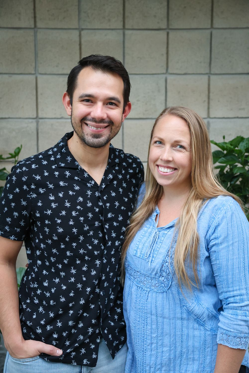 Smiling couple posing outdoors, man in black floral shirt and woman in blue blouse, standing against brick wall.
