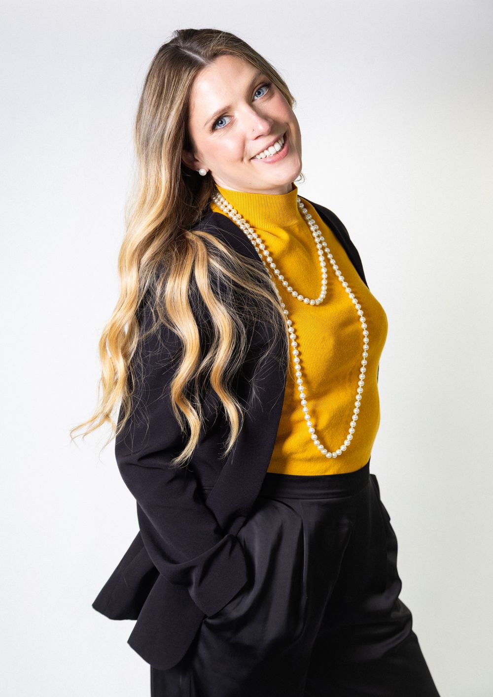 Professional studio portrait of someone wearing a mustard yellow turtleneck and black blazer with pearl necklace against white backdrop.