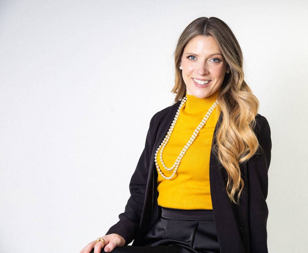 Professional portrait against white background wearing yellow turtleneck, black blazer and pearl necklace.