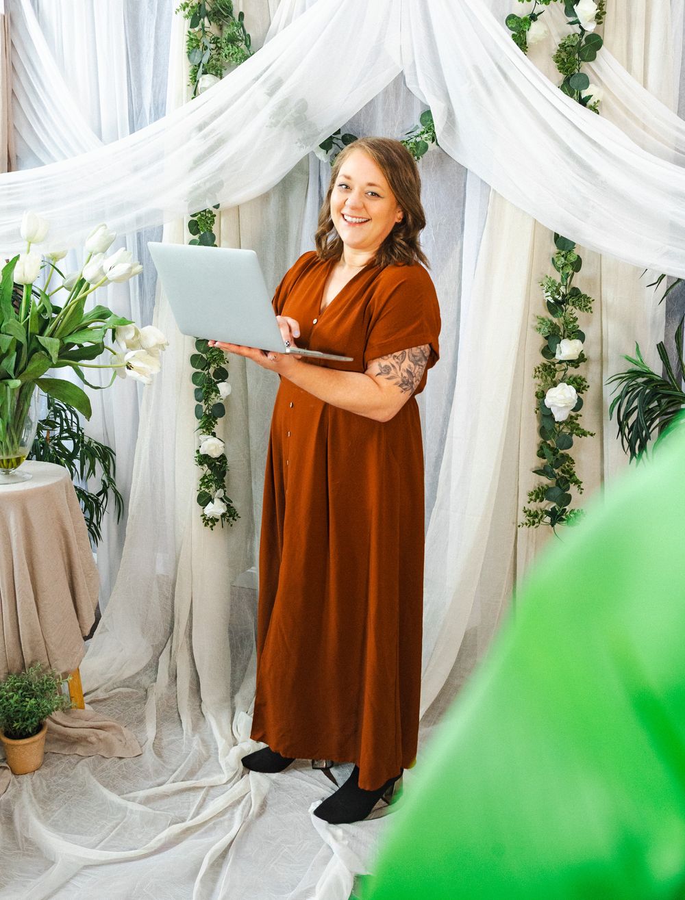 Woman in rust maxi dress holds laptop under floral arch with white draping and greenery.