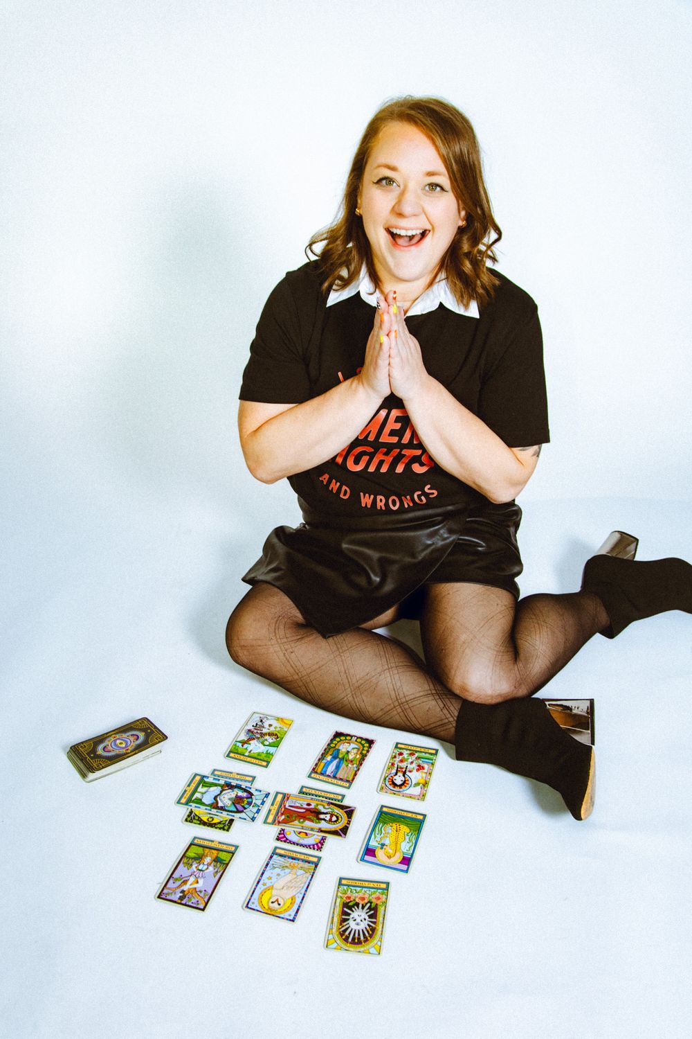Excited woman sitting cross-legged on white floor with tarot cards spread out, hands pressed together in prayer gesture.
