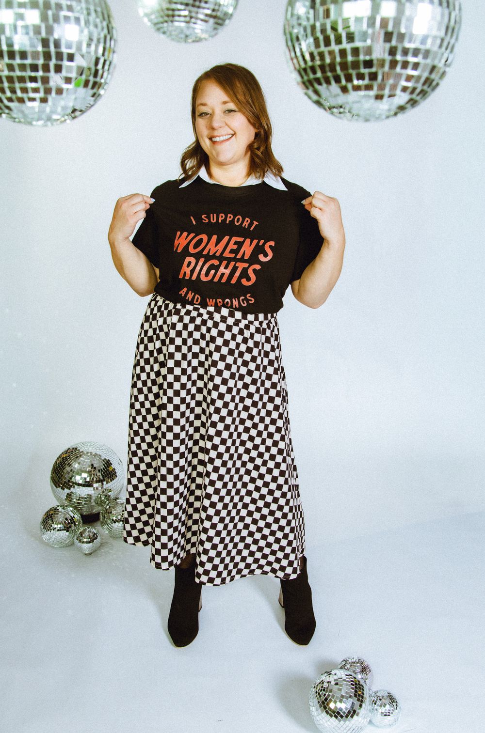 Woman in Women's Rights tee and checkered midi skirt poses with disco balls in bright studio setting.