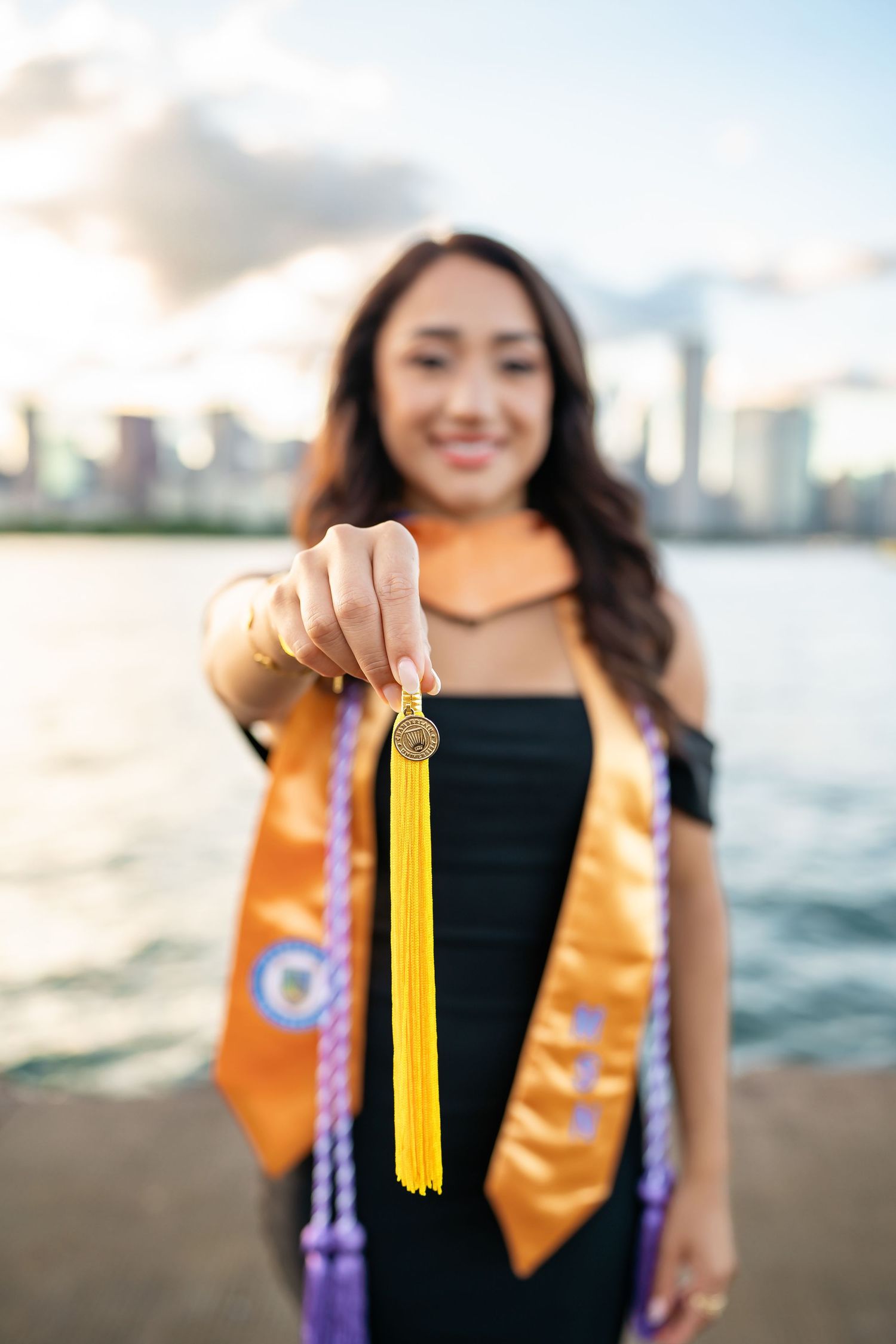 Graduate wearing black dress and orange stole holds up graduation tassels against ocean backdrop at sunset.