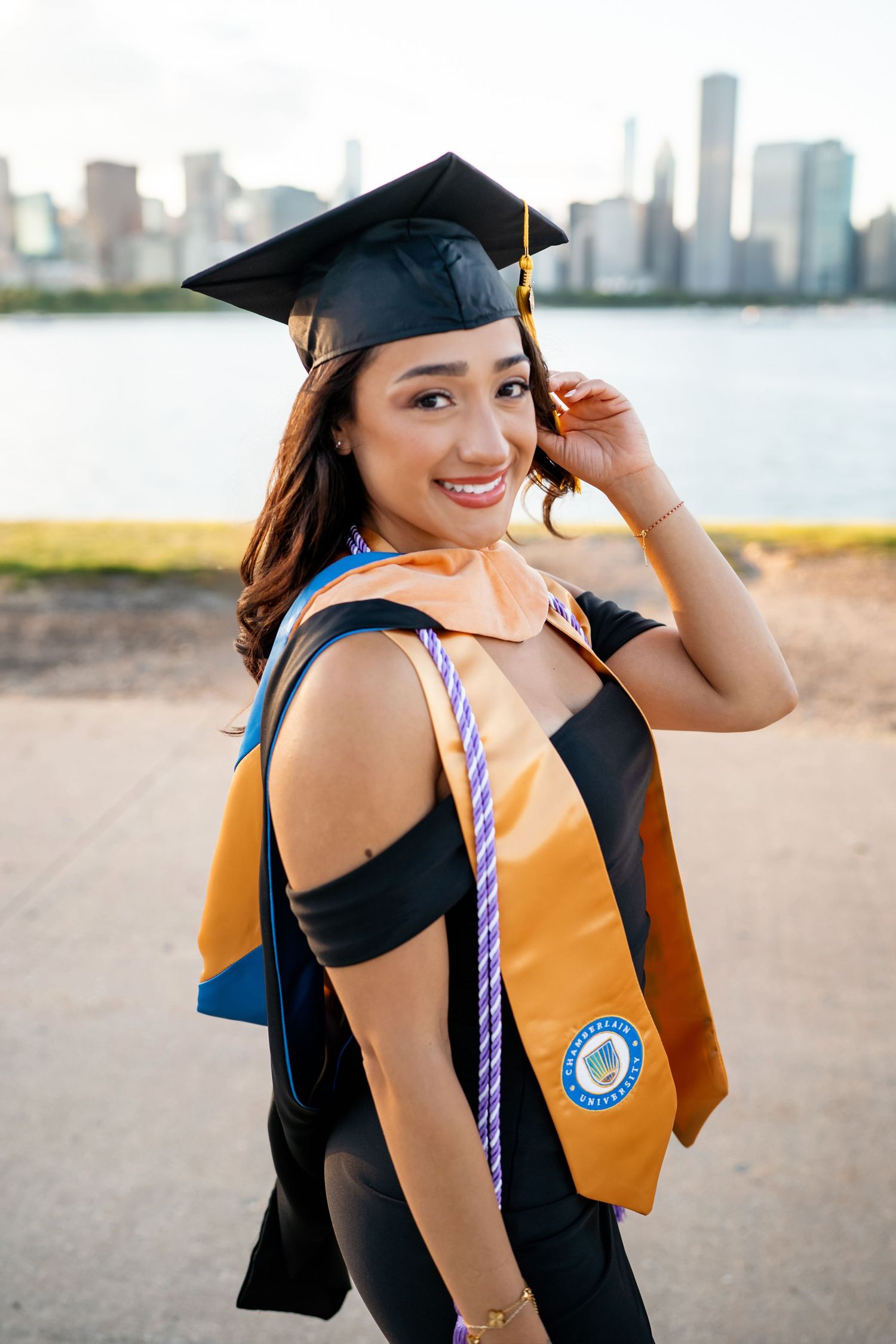 Graduate in black cap and gown with gold stole poses on beach with city skyline in background.
