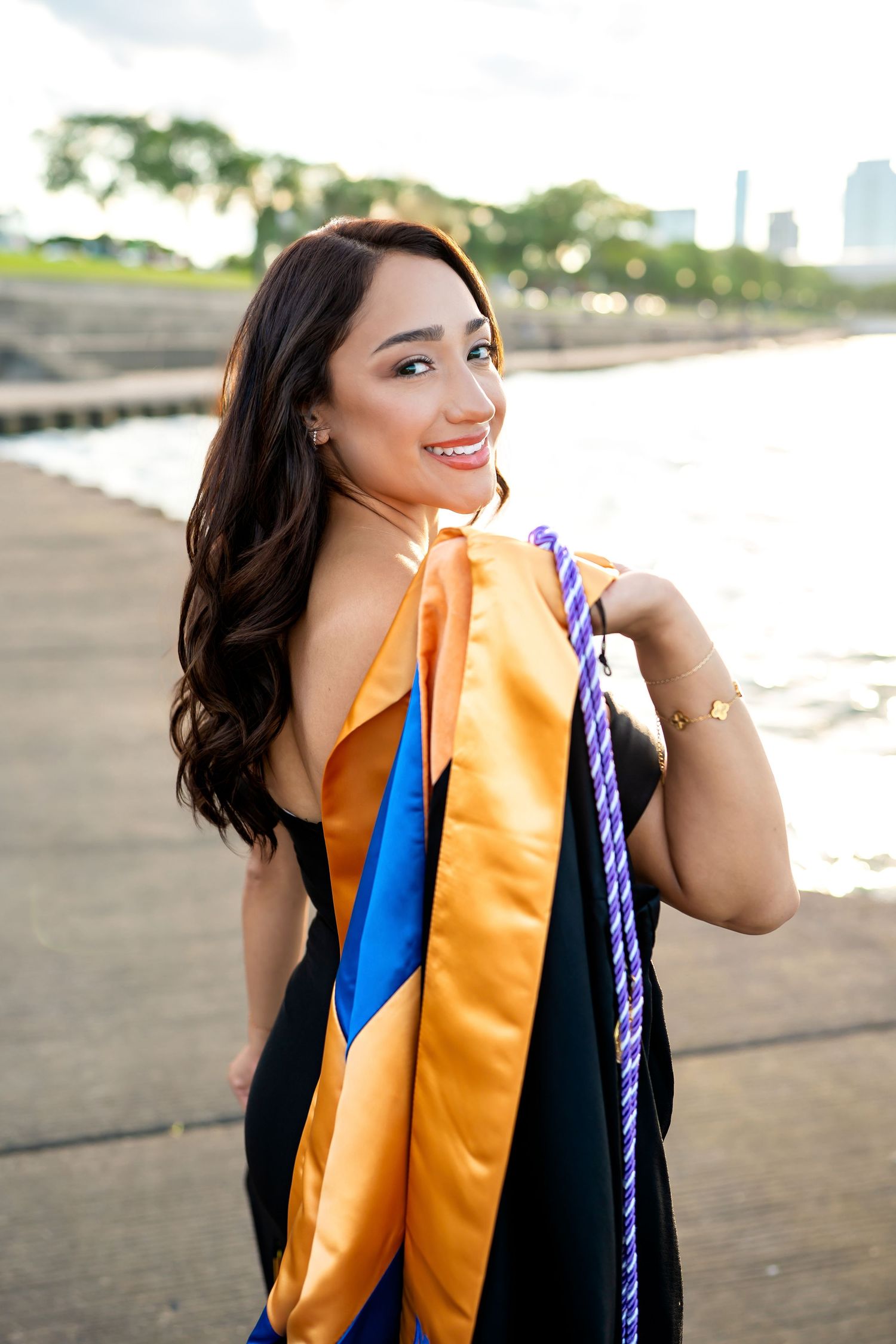 Graduate in black dress with orange and blue graduation stole smiles outdoors near waterfront with city skyline in background.