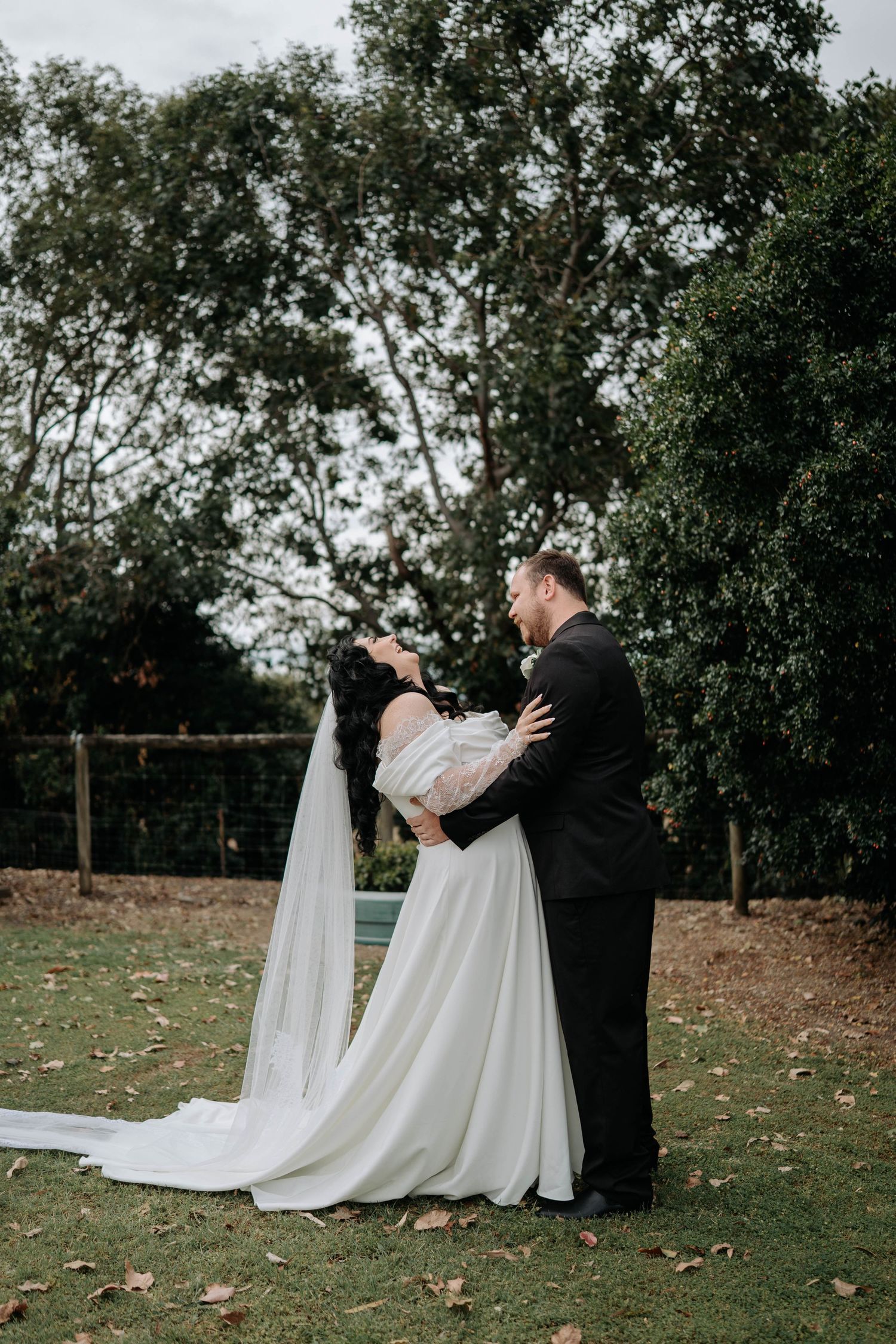 A romantic wedding couple embraces outdoors in a garden setting with flowing white dress and trailing veil.