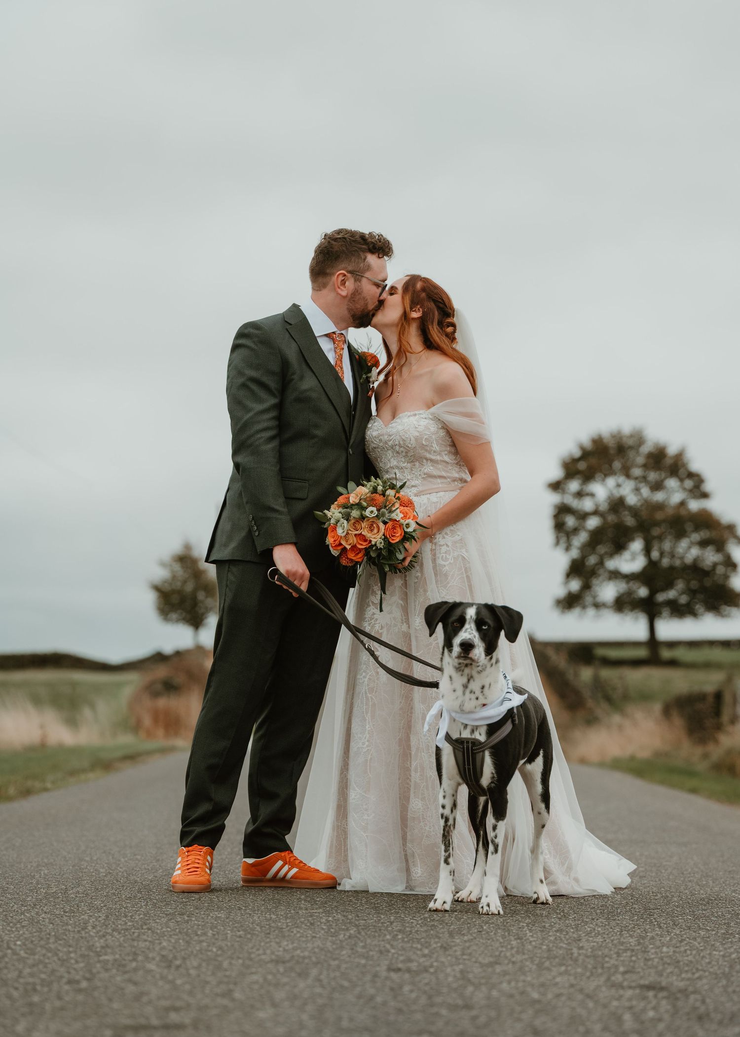 A couple shares a romantic moment on a country road with their black and white dog wearing bright orange shoes.