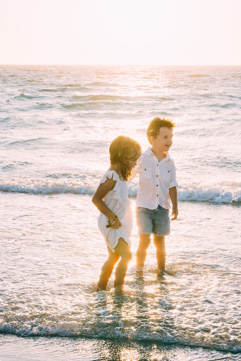 Two children play together in shallow ocean waves at sunset, creating a dreamy beach scene.