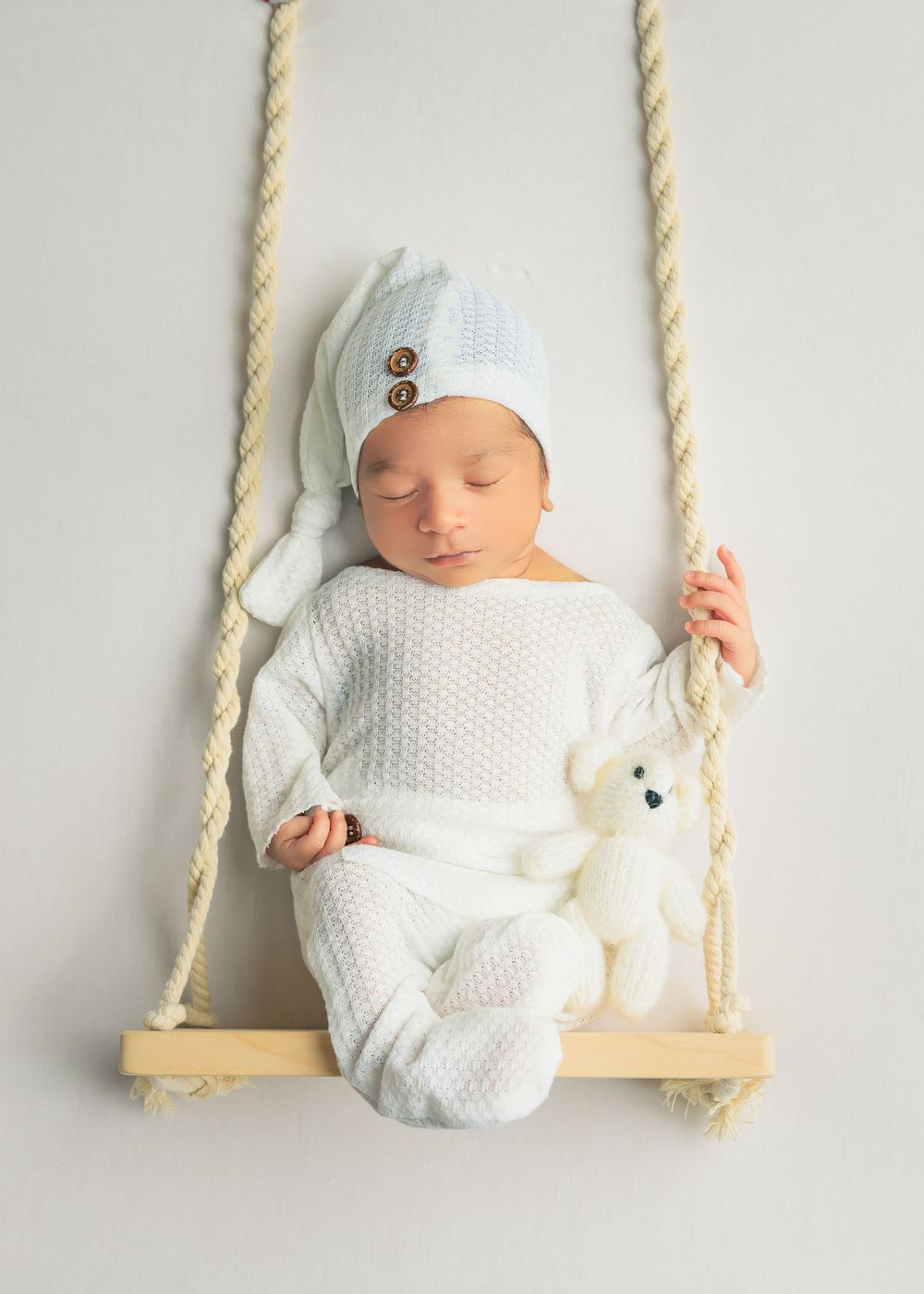 Newborn in white outfit rests on wooden swing with rope handles against white background.