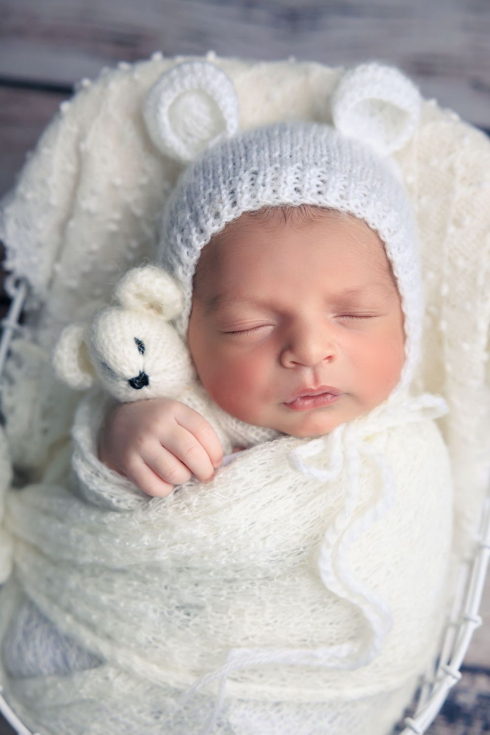 A sleeping baby wrapped in white blankets wearing a knitted hat with bear ears holds a small teddy bear.
