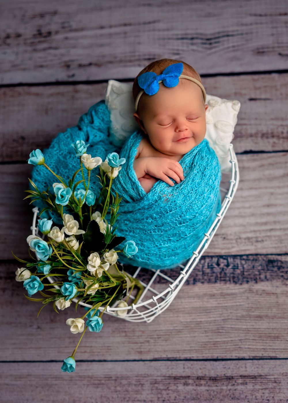 Sleeping newborn wrapped in turquoise lace with matching bow and blue roses in white basket on wooden background.
