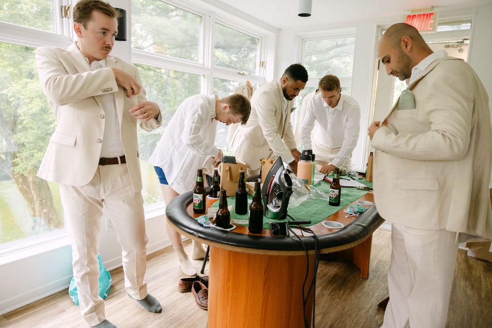 A group dressed in white suits gather around a table with wine bottles at what appears to be a pre-wedding celebration.