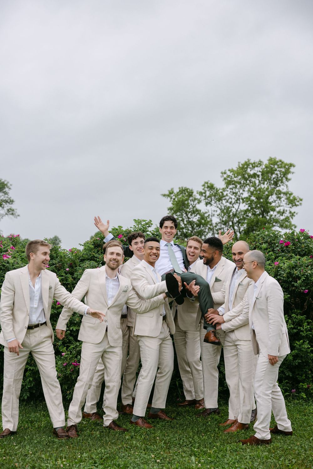 Group wearing white suits pose playfully together on a grassy hillside with green shrubs and flowers in the background.