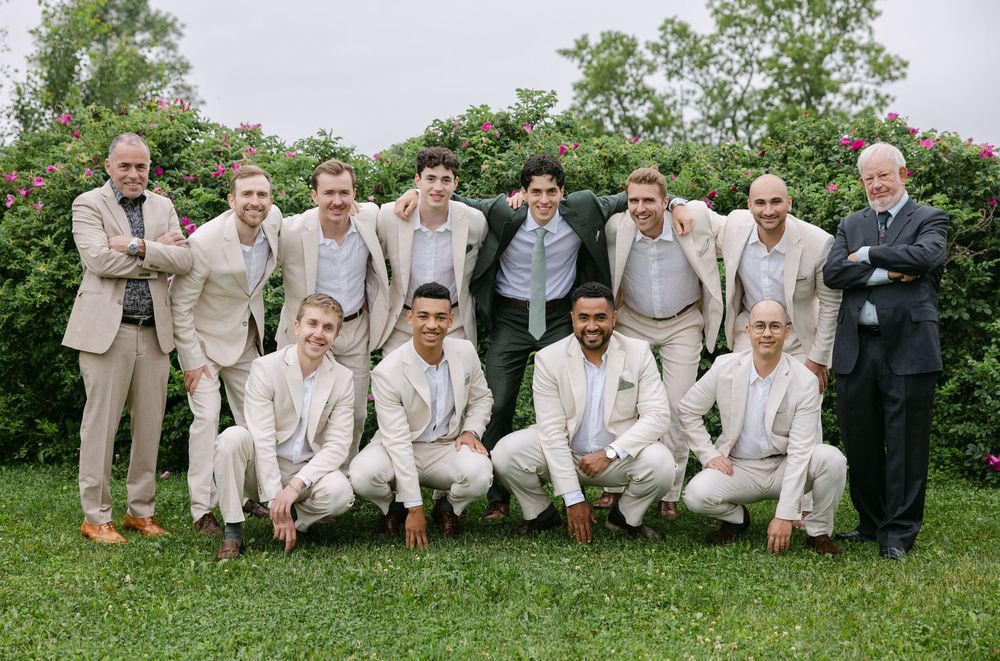 A group of people dressed in light colored suits pose together outdoors on grass with trees and bushes in the background.