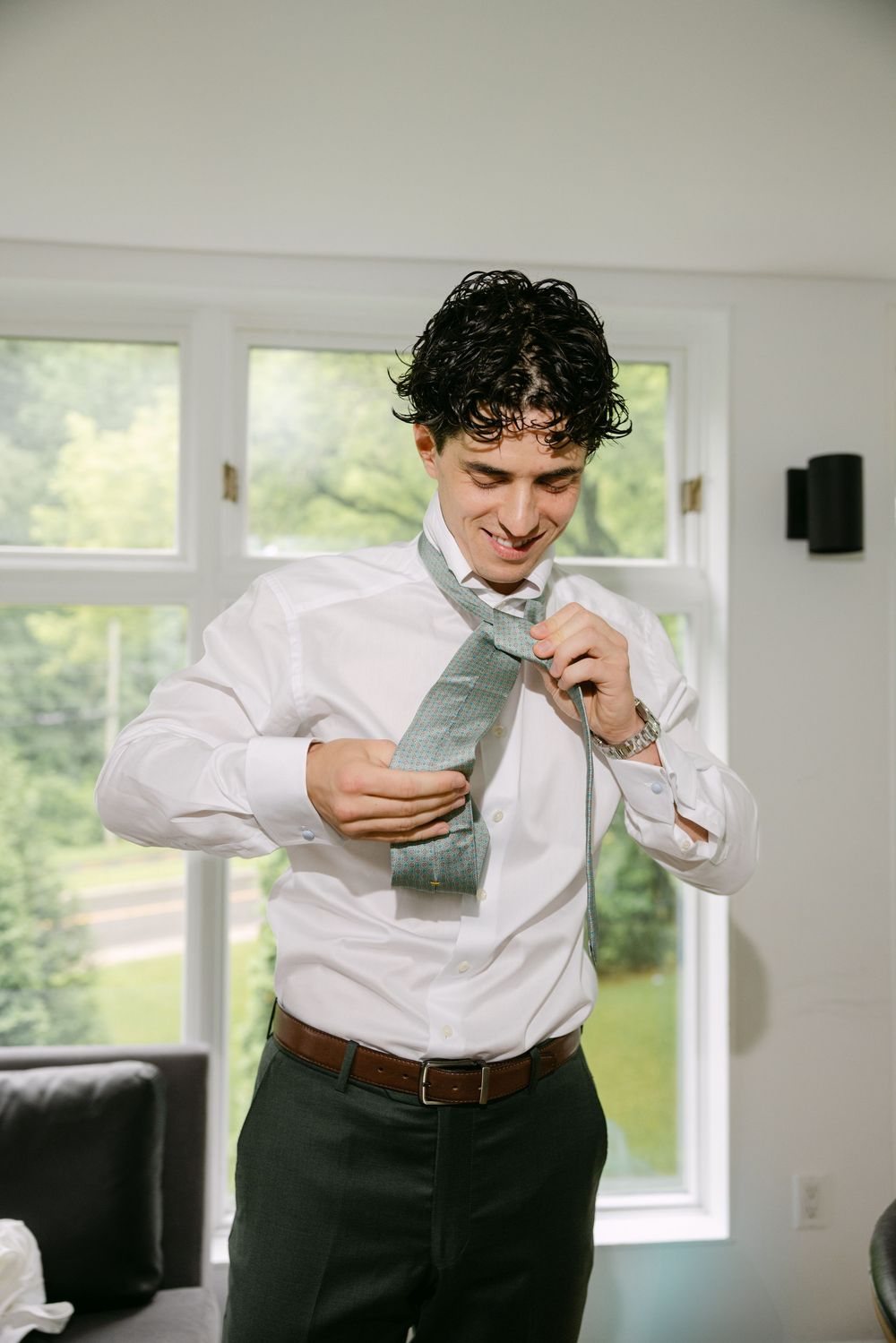 A person in business attire looks at their mobile phone while standing in a bright room with windows.