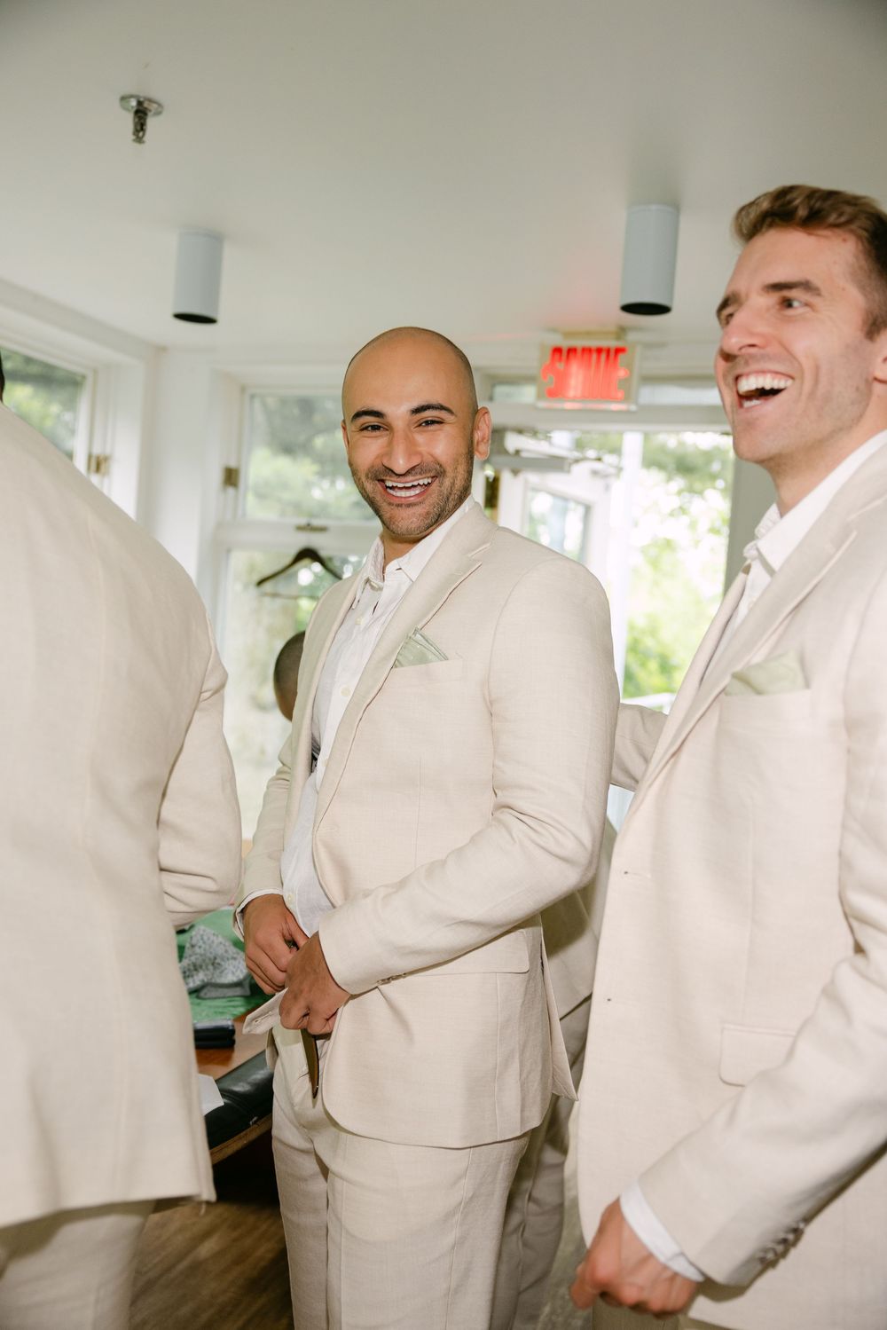 Groomsmen dressed in crisp white suits share candid laughing moments as they get ready for a wedding celebration.