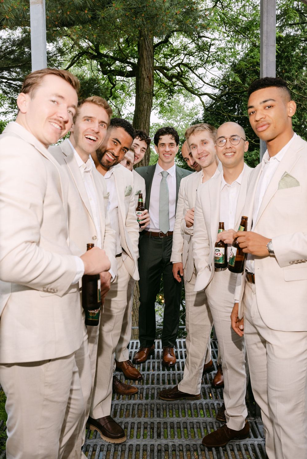 Wedding party in light colored suits gather outdoors for candid group photos against a garden backdrop.