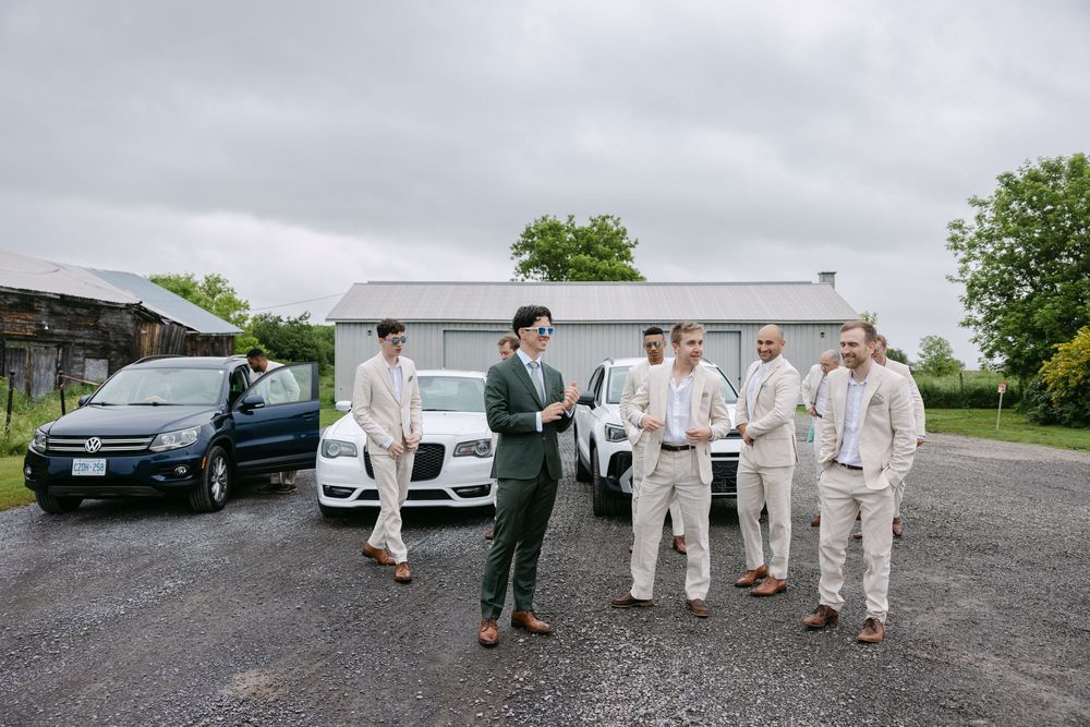 Wedding party members in beige suits gather around a white car on a gravel road on a cloudy day.