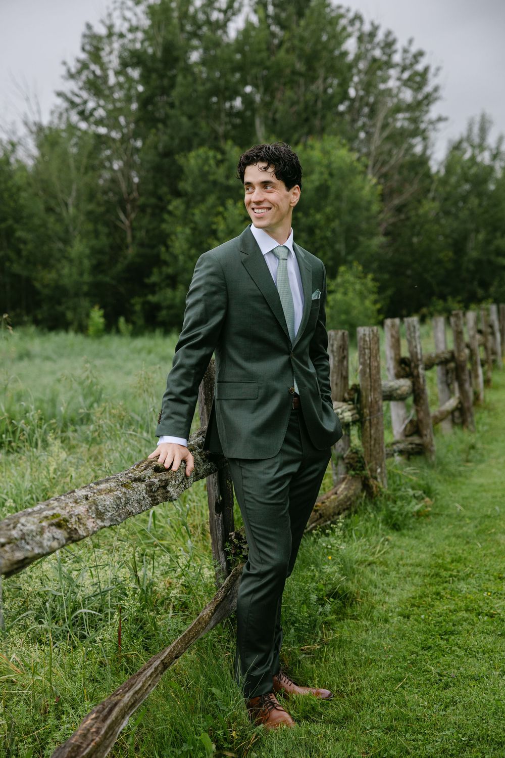 Well-dressed person in a dark green suit and blue tie stands along a rustic wooden fence in a lush countryside setting.