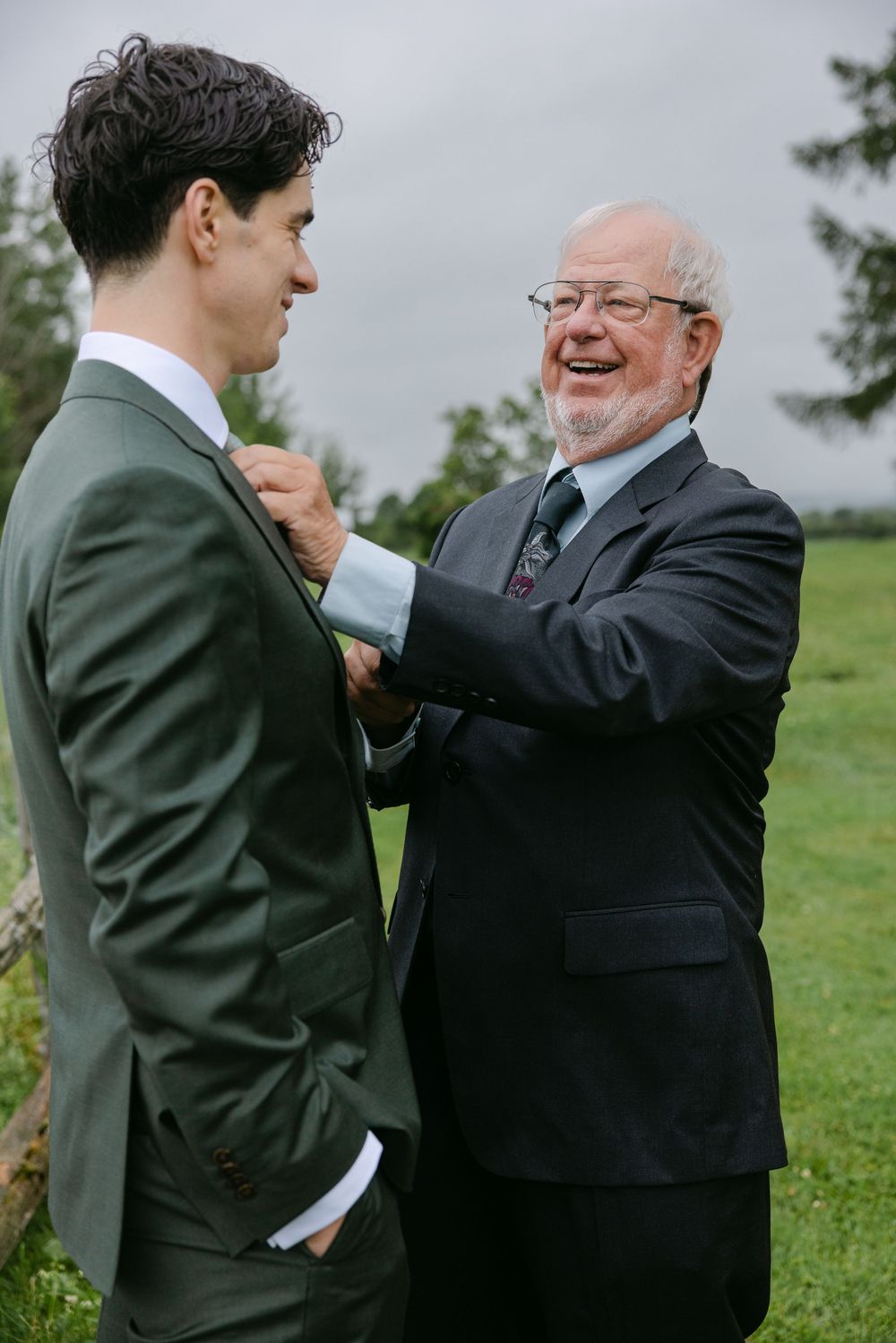Two men in suits share a joyful moment outdoors, with one helping adjust the other's suit on a grassy hillside.
