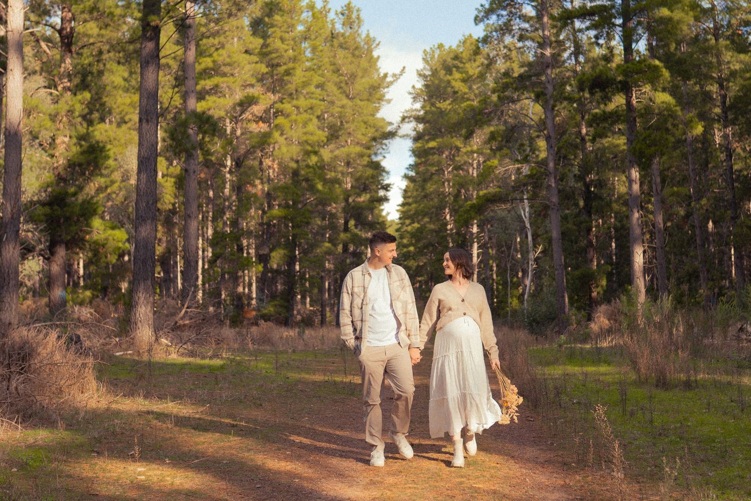 Two people hold hands while walking down a sunlit forest path surrounded by tall pine trees in the afternoon light.